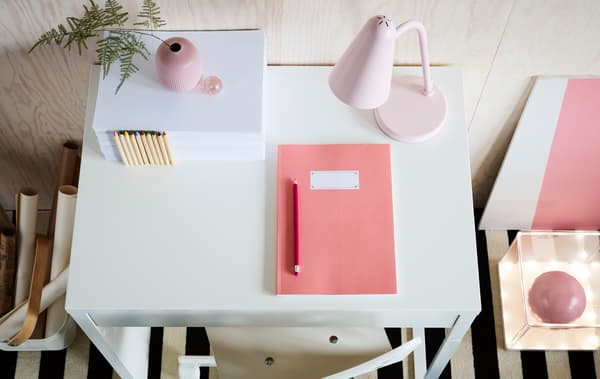  Una scrivania MICKE bianca con sopra un contenitore, un quaderno, una matita e una lampada. Il pavimento è a righe e accanto alla scrivania si trovano una scatola con all'interno dei rotoli di carta e un contenitore con delle lucine illuminate e dei pannelli rosa e bianchi. 