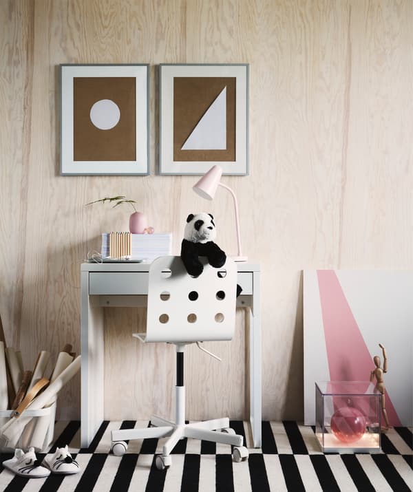 Una scrivania per bambini FLISAT in legno di IKEA al centro di una stanza con un pavimento a righe bianche e nere, due contenitori ai lati della scrivania e una sedia con un peluche di panda. 
Alla parete sono appesi due quadri raffiguranti due figure geometriche. 