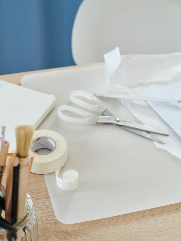 Una scrivania sopra cui sono appoggiati un un rotolo di scotch, forbici che stanno ritagliando della carta e un bicchiere con pennelli e carta.