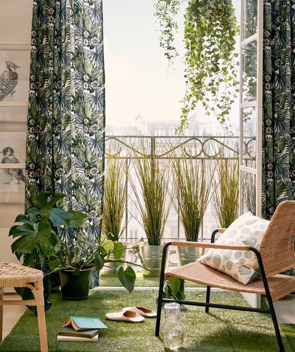 Una sedia davanti a un balcone soleggiato con la portafinestra aperta incorniciata da tende verdi, piante appese e disposte sul pavimento decorato da un prato artificiale.