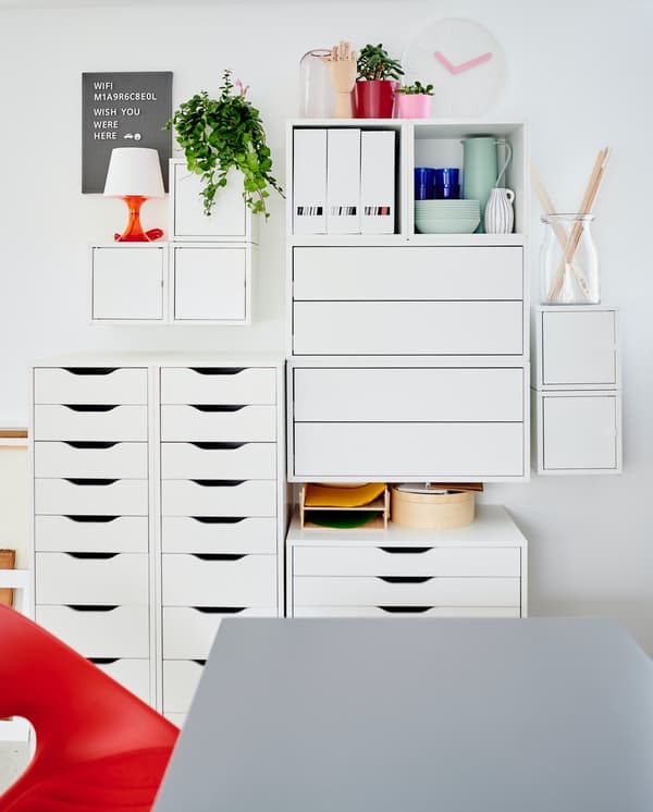 Una sedia rossa e un tavolo grigio davanti a una parete a cui sono appoggiate delle cassettiere ALEX e dei mobili da parete EKET.