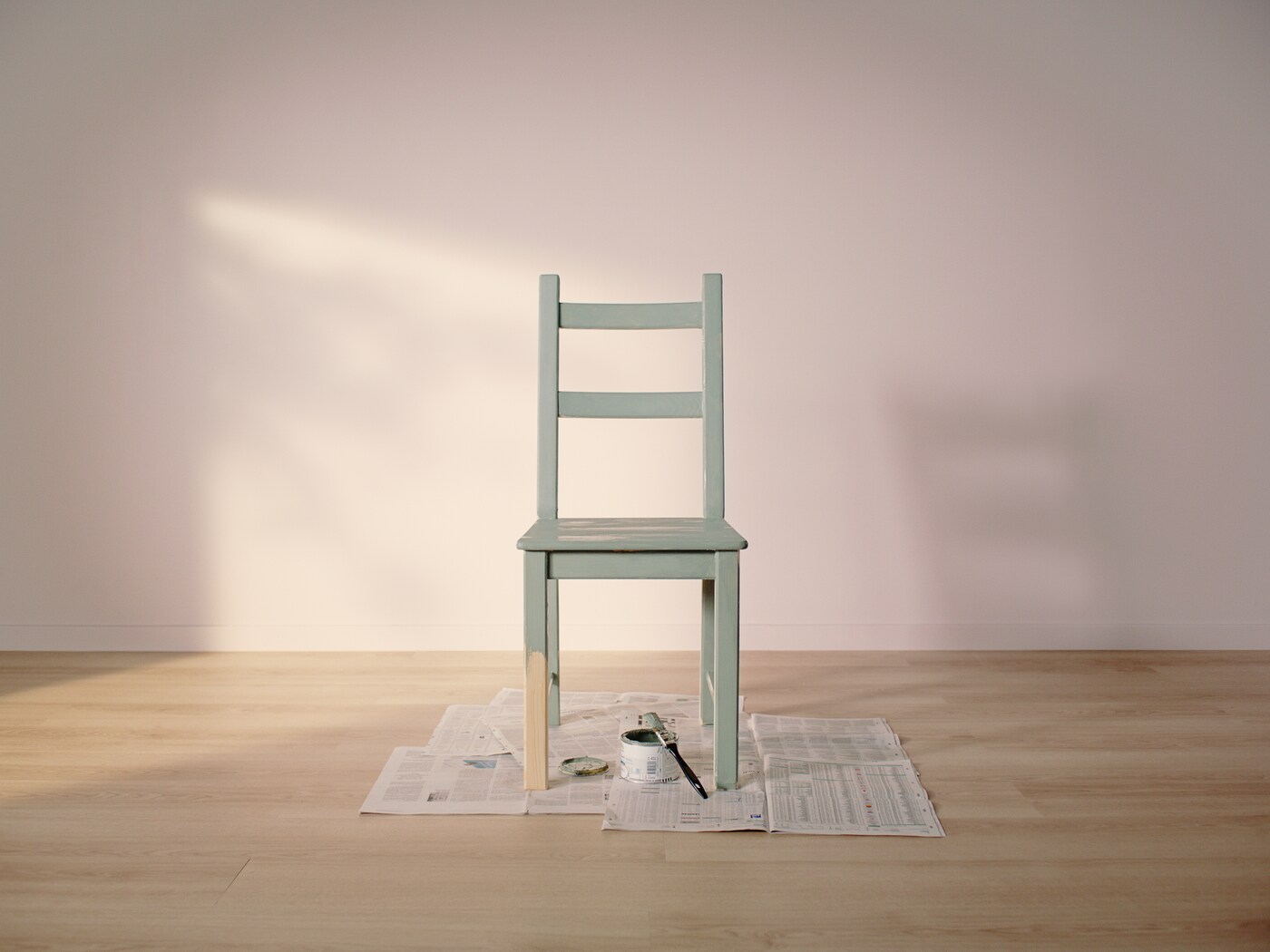 Una sedia verde menta è posizionata su un pavimento in legno, circondata da fogli di giornale. La luce del sole proietta ombre morbide sulla parete, creando un'atmosfera calma e minimalista.