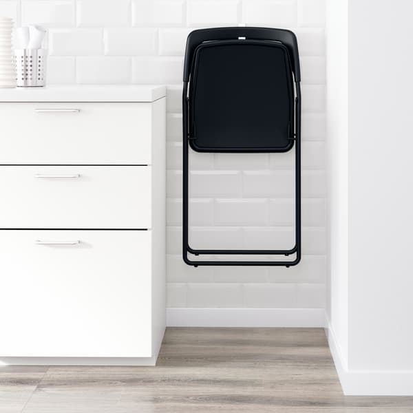 Una silla plegable negra IKEA NISSE colgando de una pared blanca en un espacio estrecho entre una pared y un armario.