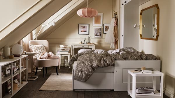 Una stanza con un letto divano bianco aperto con biancheria da letto beige/grigio scuro, vari mobili, una poltrona beige e una lampada a sospensione rosa - IKEA
