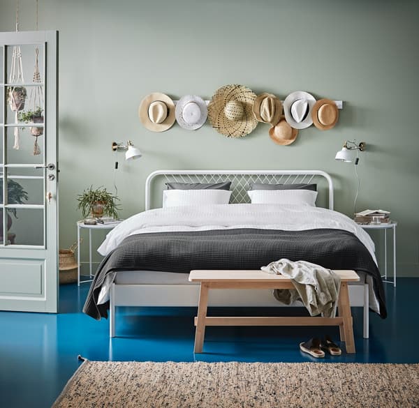 Una stanza con un parete verde chiari e un pavimento azzurro. Al centro della stanza un letto e un tavolino con sopra degli abiti e sotto delle scarpe. Accanro al letto due tavolini con sopra una piante e un oggetto. 
Sulla parete sono appesi dei cappelli di paglia.