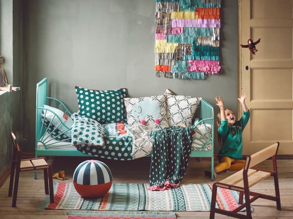Una stanza per bambini giocosa in stile Boho con un letto verde sulla parete in fondo coperto con molti tessuti. Piccoli tappeti e peluche giacciono sul pavimento.