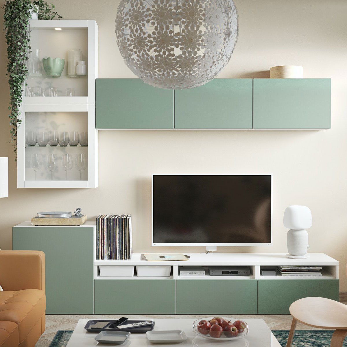 Una struttura bianca e verde BESTÅ in un salotto in una composizione con strutture ad anta in vetro e anta colorata. La TV è agganciata a parete e si intravede un divano, una poltrona e un tavolino. In alto, si vede una lampada a sospensione GRIMSÅS.
