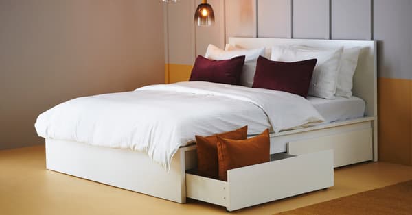 Una struttura letto alta con 2 contenitori bianca MALM. Le lenzuola sono bianche e i cuscini hanno federe marroni.