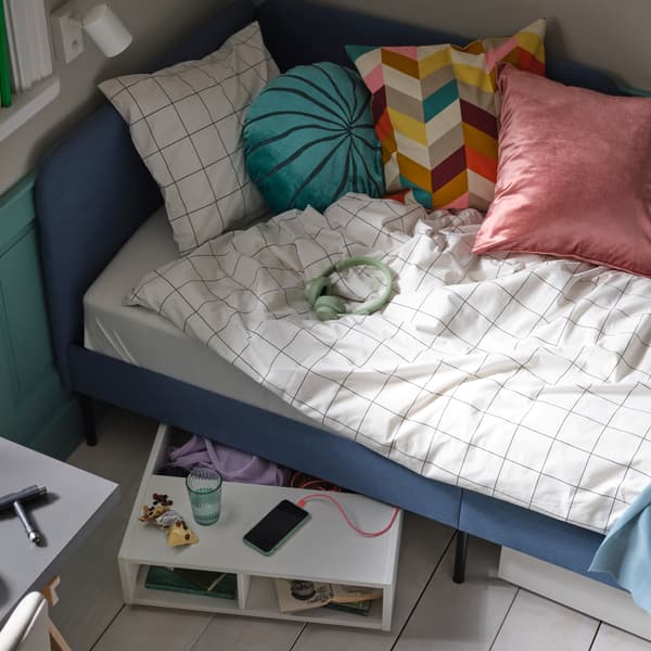 Una struttura letto blu imbottita BLÅKULLEN , con biancheria da letto VITKLÖVER bianca e nera a quadri e vari cuscini, più un contenitore sottoletto FREDVANG.