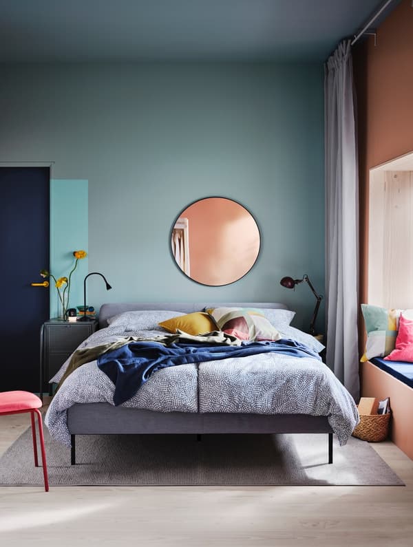 Una struttura letto imbottita SLATTUM in una camera da letto con pareti azzurre, una porta blu, un vaso con fiori nero, una lampada da tavolo nero e una sedia rossa.  