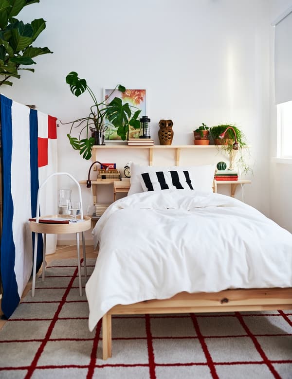 Una struttura letto NEIDEN con affianco un tavolino, dietro due mensole con sopra delle piante e dei libri. Accanto al tavolino una parete con fantasie a strisce e quadrati.