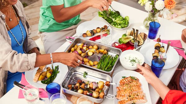 Una tavola all'esterno su cui sta venendo consumato un pranzo abbona con verdure, carne e pesce.  Al tavolo ci sono 4 persone.