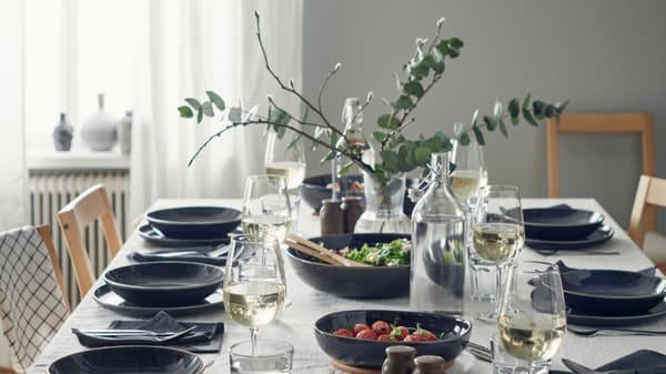 Una tavola allestita con una tovaglia bianca, bicchieri vino bianco STORSINT, bottiglie d'acqua in vetro KORKEN, bicchieri POKAL, servizio grigio scuro opaco FÄRGKLAR e saliere/pepiere BRUGDHAJ. Al centro, dell'insalata e un vaso trasparente PÅDRAG con dei rami di eucalipto.
