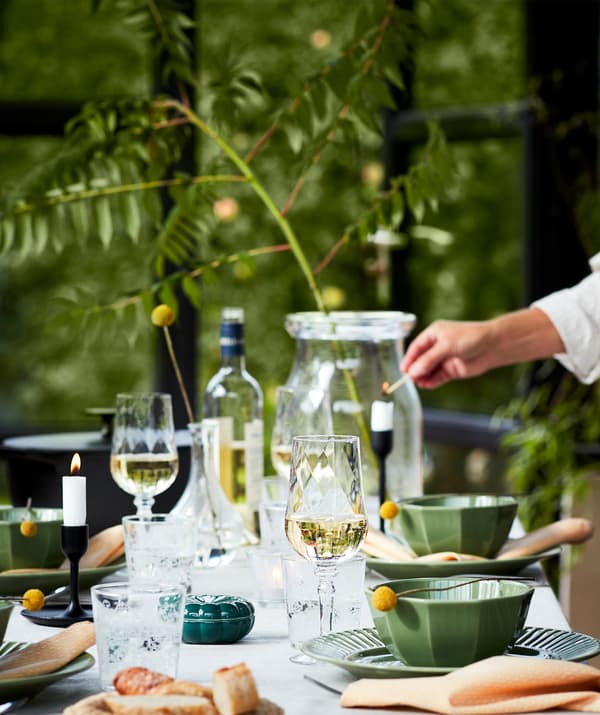 Una tavola apparecchiata per una cena all’aperto con piatti verdi, bicchieri da vino, candele e altri accessori, in una cornice naturale.