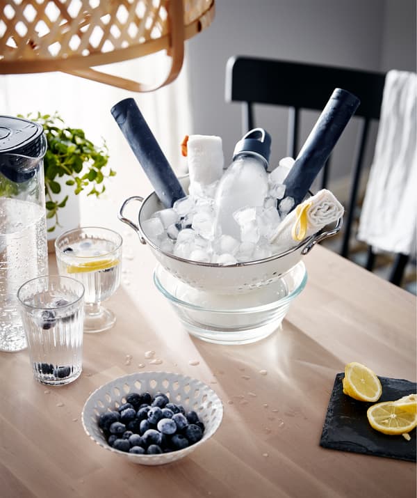 Una tavola con alcuni elementi rinfrescanti, come dell'acqua ghiacciata nei bicchieri e in una brocca, e un colapasta pieno di ghiaccio sopra una ciotola - IKEA