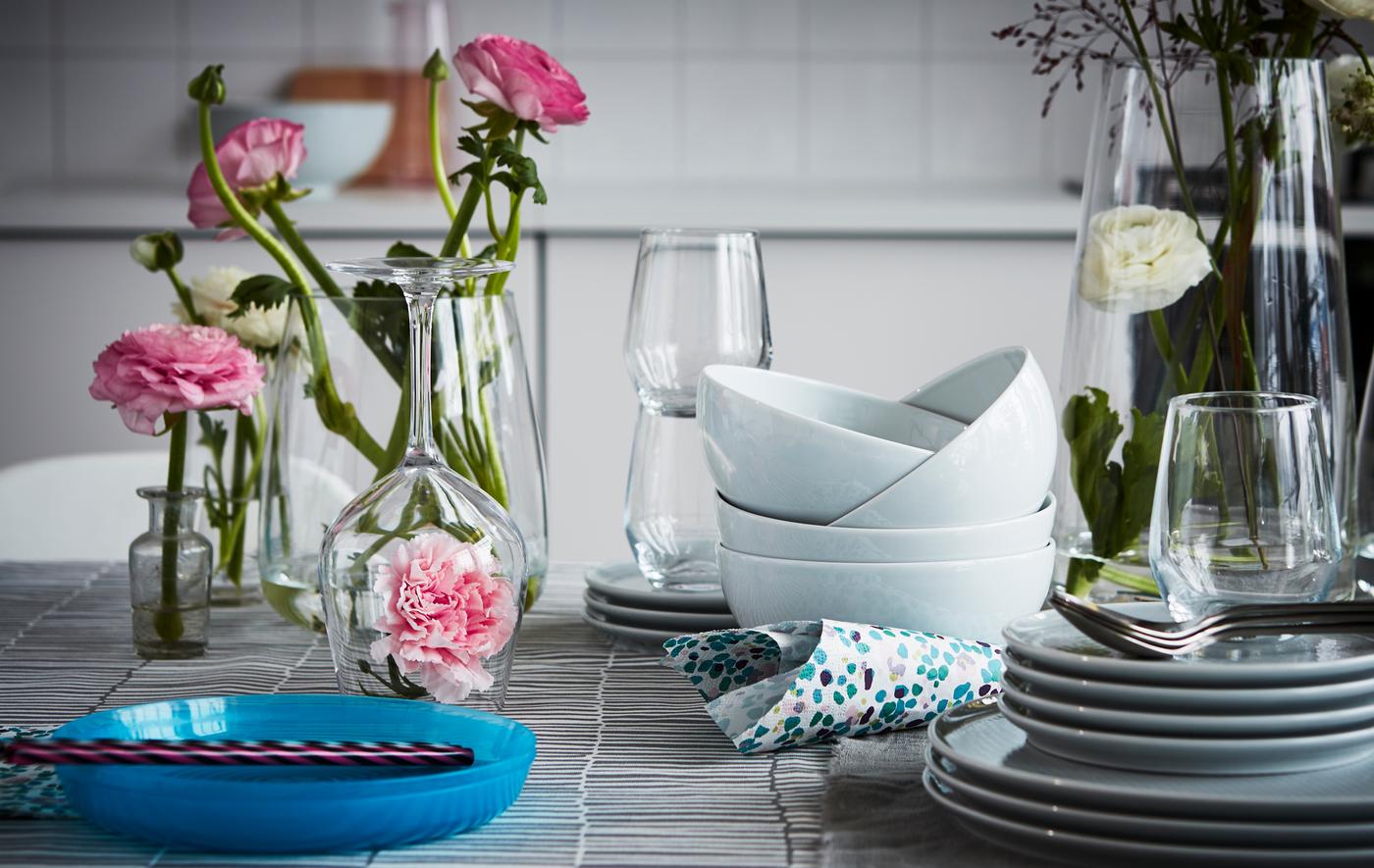 Una tavola con posate, accessori, bicchieri da vino bianco IVRIG in vetro trasparente e ciotole. Ci sono anche vasi con fiori e un fiore sotto un bicchiere da vino in vetro.