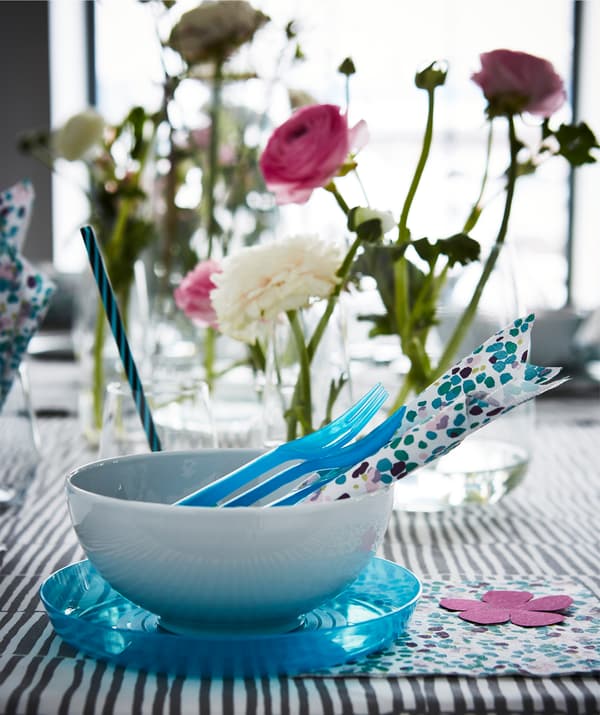 Una tavola con posate, accessori, bicchieri e ciotole bianchi e azzurri. Ci sono anche vasi con fiori.