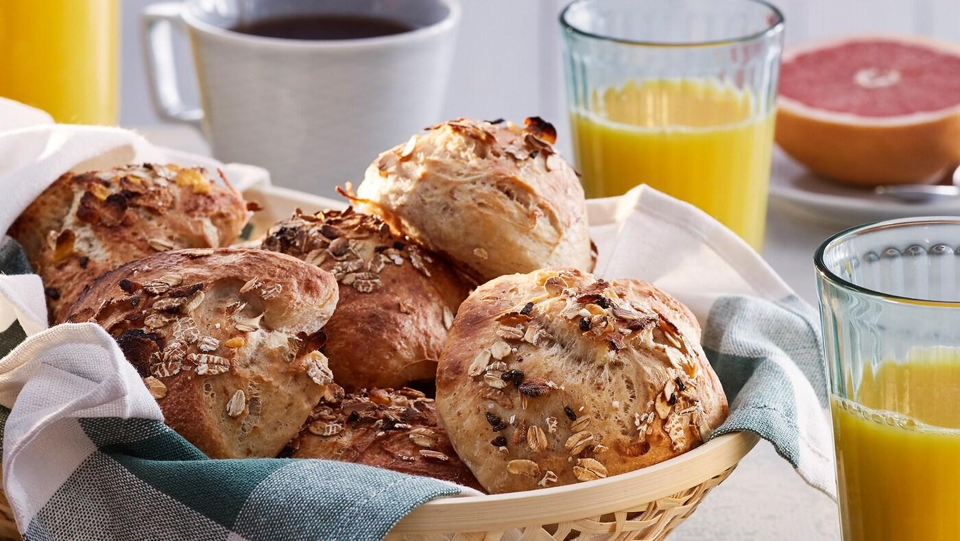 Una tavola con un cestino di panini con muesli e fiocchi di sale. Accanto, bicchieri con spremute di agrumi e tazze di caffè.
