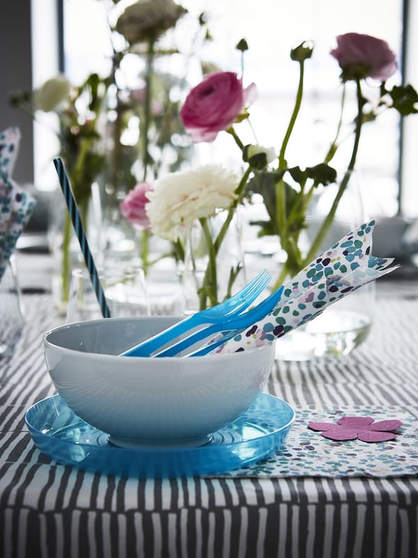 Una tavola con una ciotola, alcun posate e tovaglioli. Sullo sfondo, ci sono vasi trasparenti con fiori.