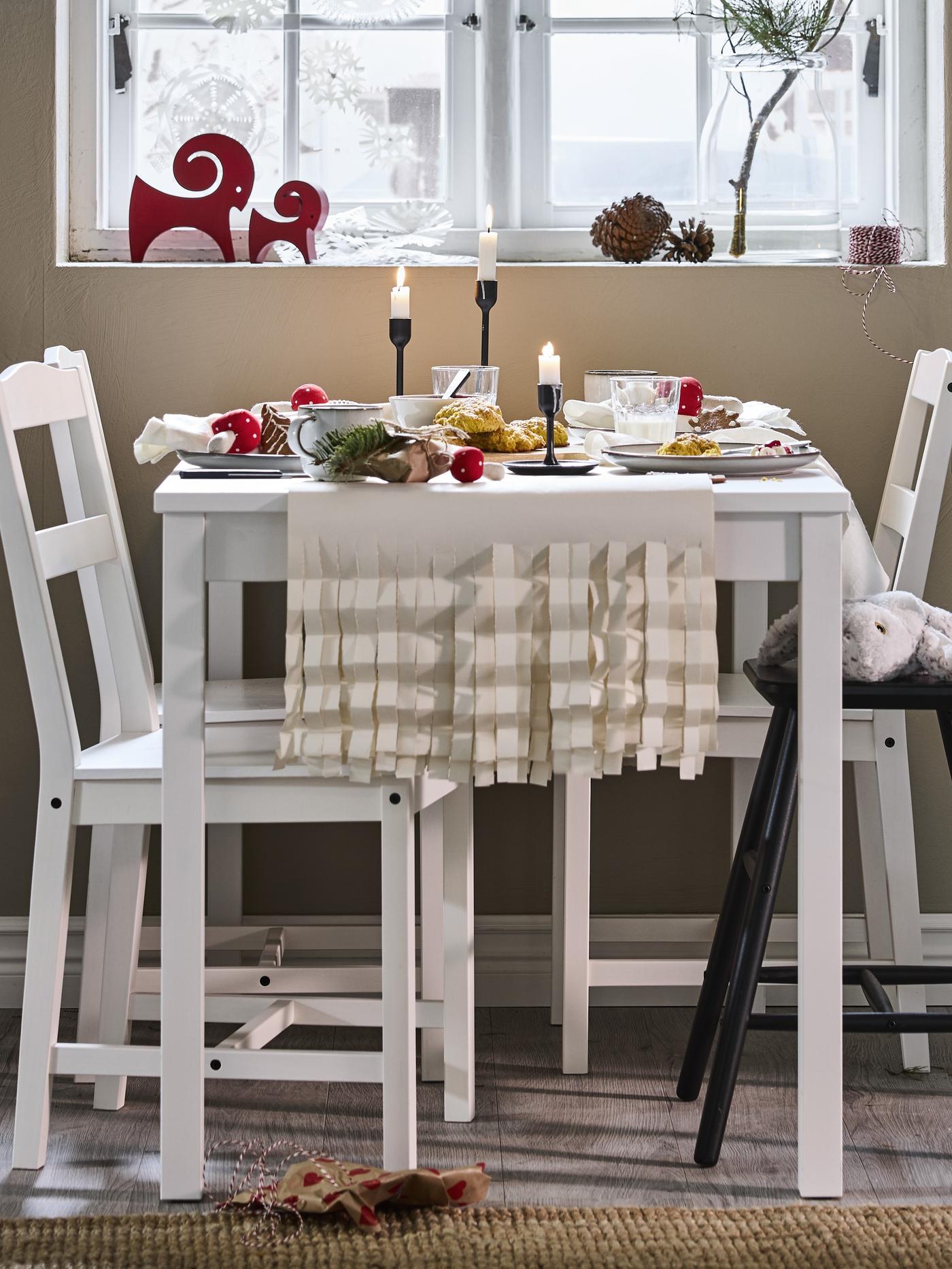 Una tavola e sedie bianche allestita per Natale con candelieri FULLTALIG con candele accese. Dietro, un davanzale con decorazioni a tema.