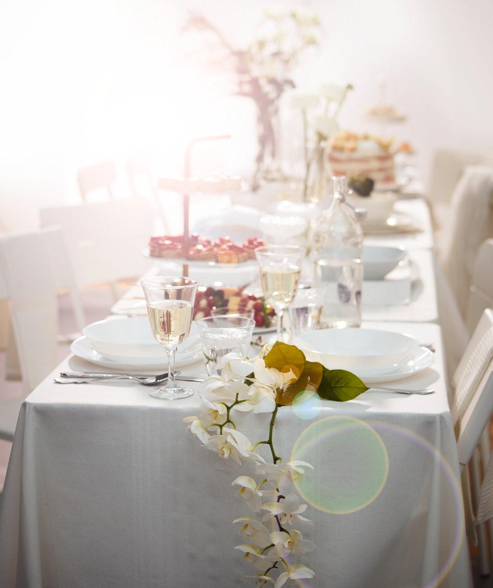 Una tavola sui toni del bianco allestita per un matrimonio. Ci sono fiori, alzate, bottiglie e bicchieri di vetro per l'acqua e per il vino illuminati da una luce.