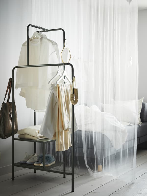 Una tenda bianca sottile separa un divano da uno stand appendiabiti NIKKEBY grigio-verde con scarpe, vestiti e accessori.