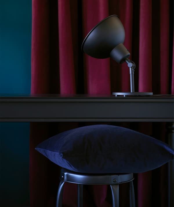 Una tenda color bordeau. Davanti, una scrivania con lampada e sedia in legno nero d'ispirazione industriale.