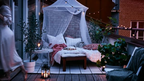 Una terrazza al tramonto con un angolo accogliente arredato con un lettino, un baldacchino a rete, un tappeto, lanterne e fili di luci da esterno - IKEA
