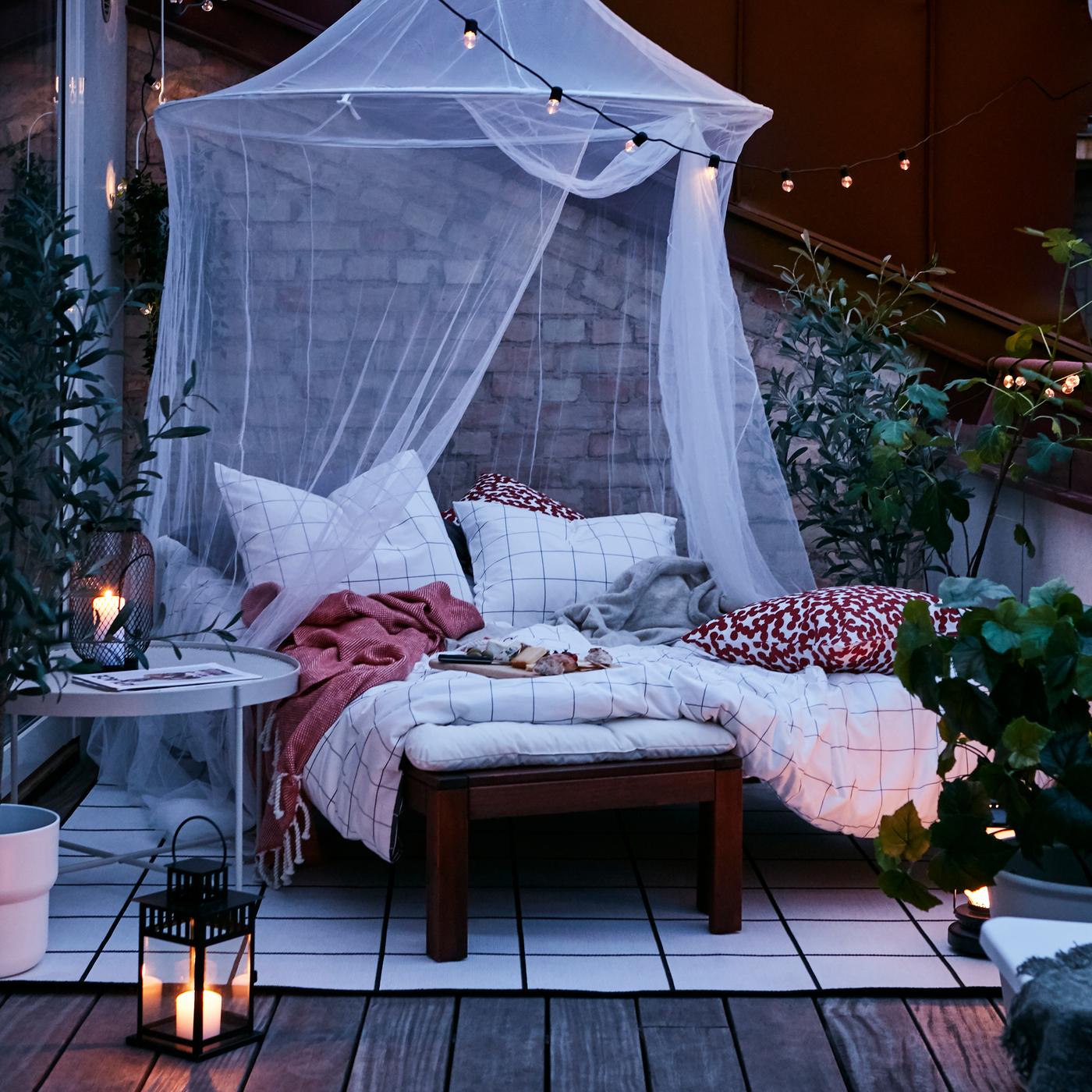 Una zona al aire libre con un camastro café preparado como una cama con ropa de cama a cuadros en blanco y negro sobre una alfombra bajo una mosquitera blanca.