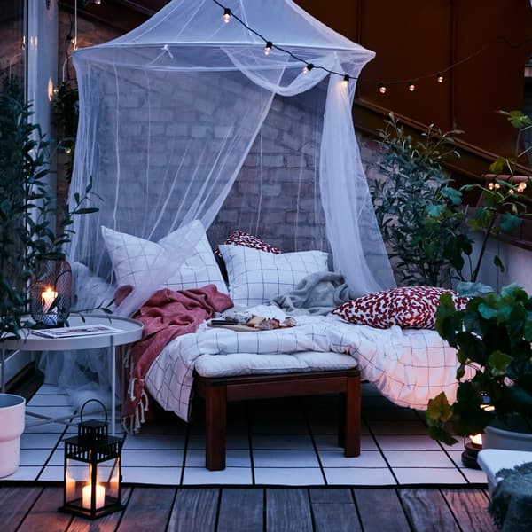 Una zona al aire libre con una tumbona marrón preparada como una cama con ropa de cama a cuadros en blanco y negro sobre una alfombra bajo una mosquitera blanca.