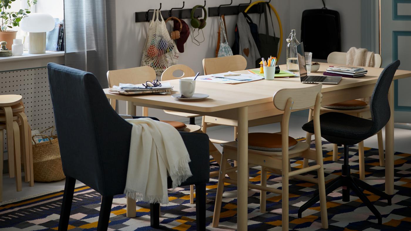 Una zona de comedor con una mesa de abedul RÖNNINGE con varios objetos encima y sillas RÖNNINGE sobre una alfombra TÅRBÄK.