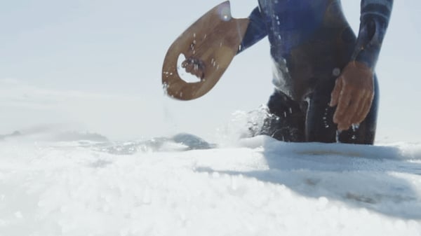 Underkropp står i vitt skum från havet och håller i en liten bräda i trä