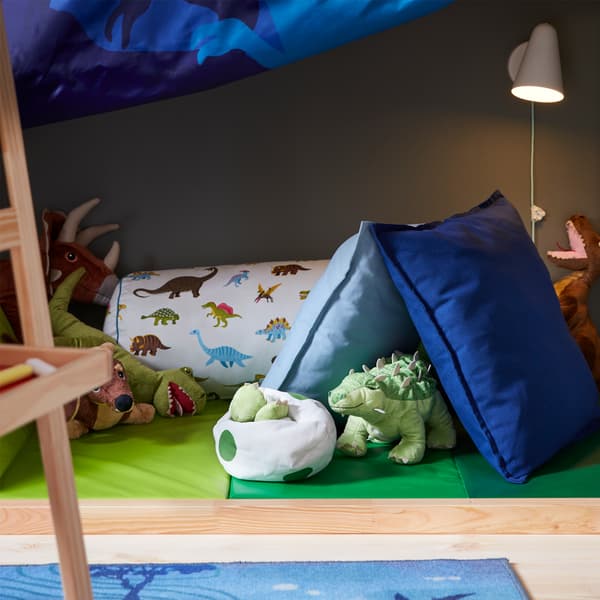 Une applique blanche allumée et un tapis de gymnastique pliant vert sur lequel sont posés des dinosaures en peluche et des coussins bleus/à motifs de dinosaure.