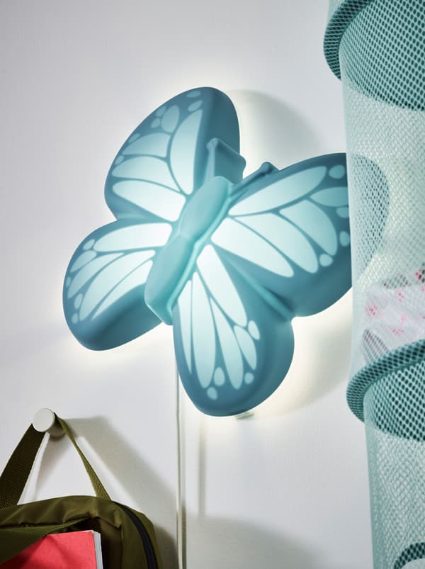 Une applique LED UPPLYST turquoise en forme de papillon placée entre un sac suspendu à un crochet et un tube de rangement en filet.
