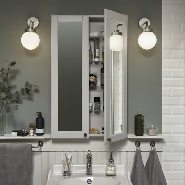 Une armoire à miroir, haute et peu profonde, au-dessus d’un lavabo blanc. Une porte ouverte, articles de toilette visibles à l’intérieur.