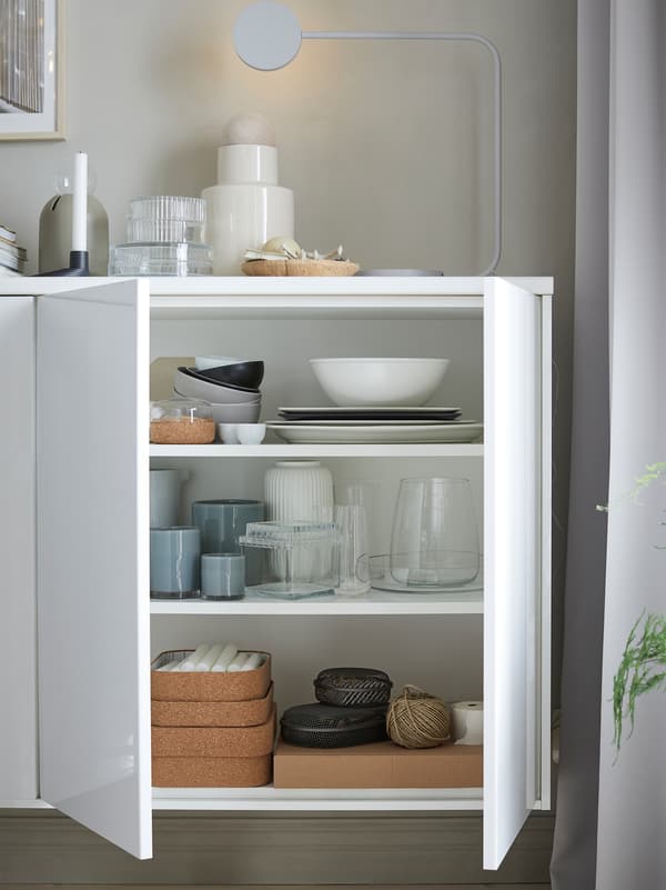 Une armoire blanche avec des portes blanc brillant ouvertes révélant un service de table, des vases et des bougies. Une lampe est posée dessus.