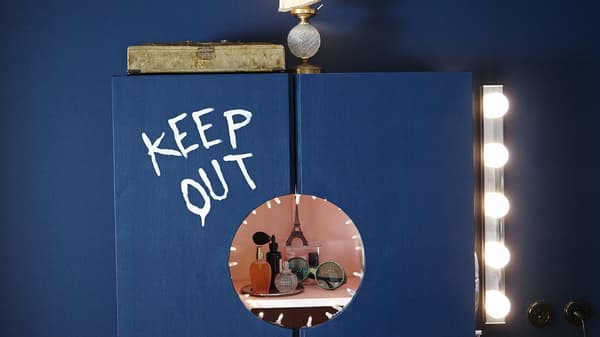 Une armoire bleue sur laquelle sont peints les mots « Keep out ». Un cercle a été découpé pour révéler des flacons de parfum et des lunettes de soleil.