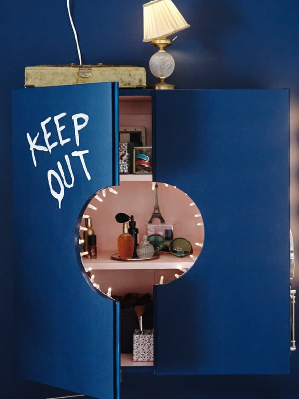 Une armoire IVAR peinte en bleu entrouverte, révélant des tablettes et un fond roses, est remplie de produits de beauté et d’accessoires.