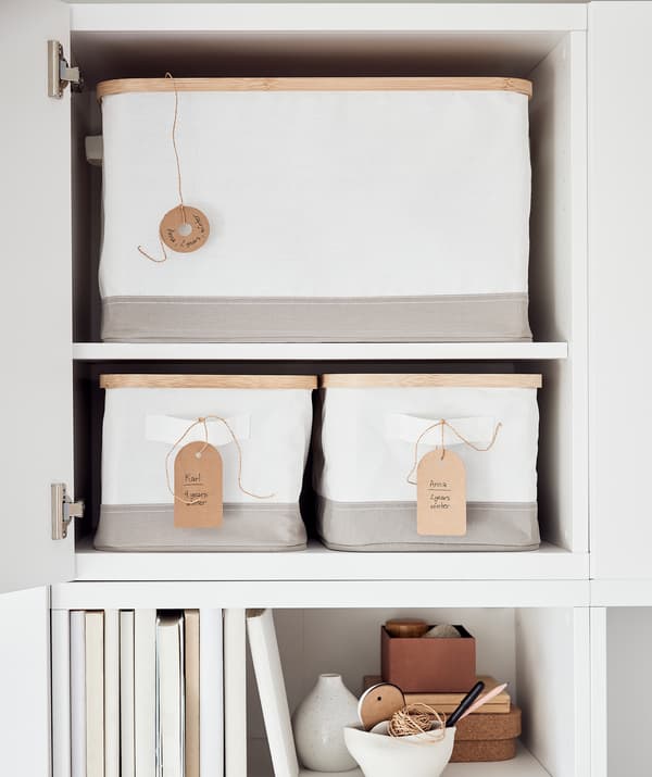 Une armoire, la porte ouverte, dévoilant des boîtes de rangement RABBLA portant des étiquettes écrites à la main. Sous l’armoire, une étagère avec des livres.