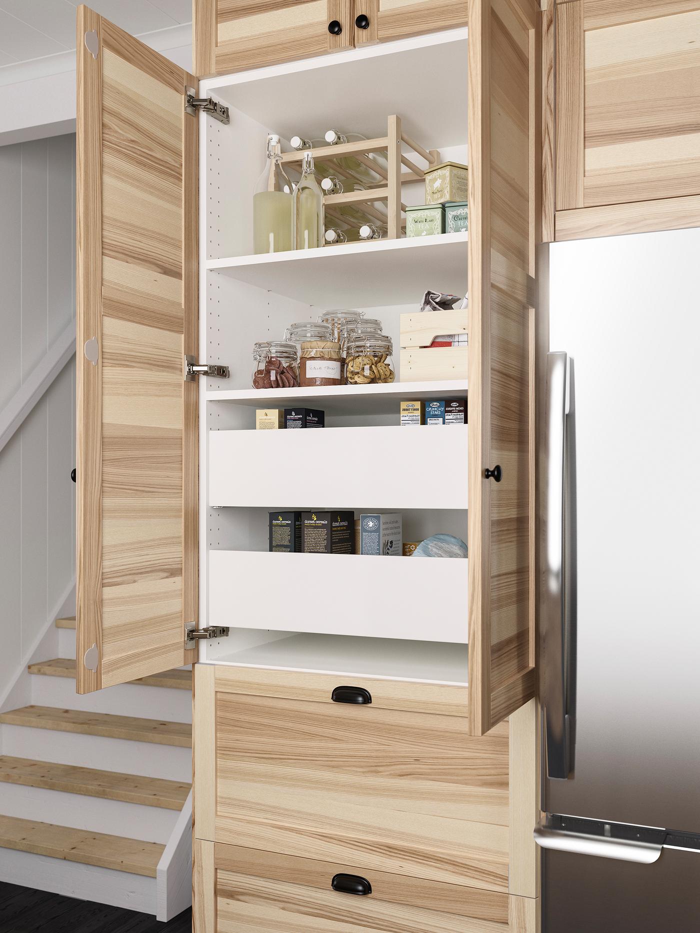 Une armoire murale ouverte avec des portes en frêne clair et, à l’intérieur, deux tiroirs blancs et des tablettes où sont rangés des aliments secs et de la limonade.