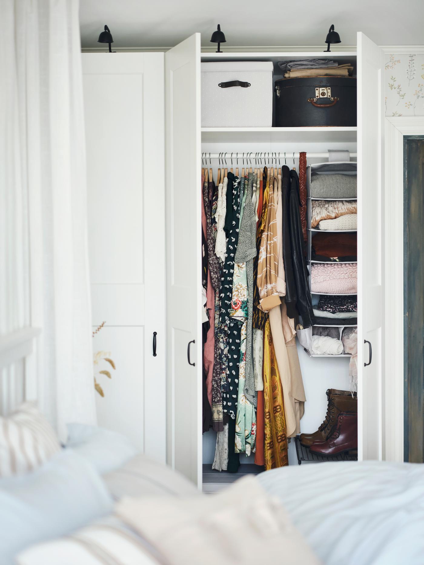 Une armoire-penderie PAX/GRIMO devant un lit double, avec deux portes ouvertes montrant des vêtements pendus et pliés et des boîtes de rangement.