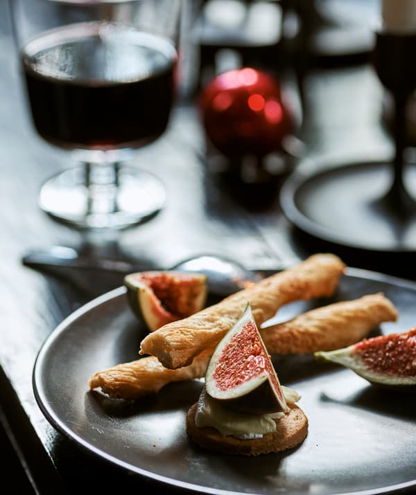 Une assiette avec de la nourriture festive: des figues, du fromage, des biscuits et des gressins, sur une table avec un verre de vin et une boule de Noël rouge.