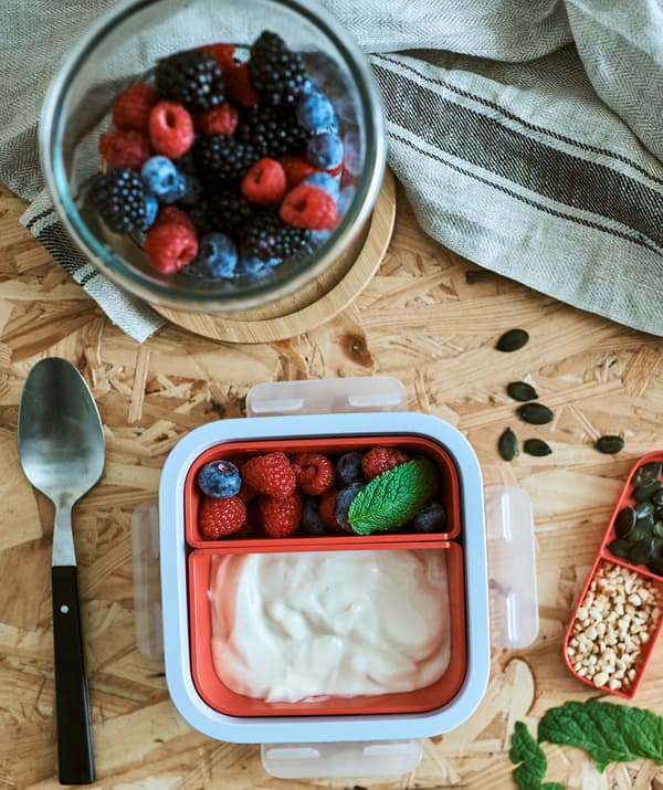 Une boîte à déjeuner blanche avec des inserts de couleur orange, l’un contient du yaourt, l’autre des fruits rouges, à côté se trouve un bocal en verre rond avec quelques baies.