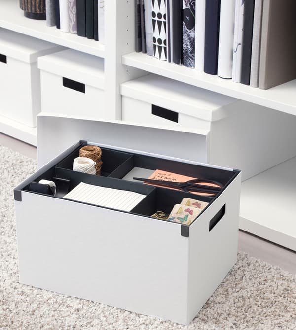 Une boîte de rangement blanche TJENA, posée sur un tapis gris clair, avec d’autres boîtes en arrière-plan.