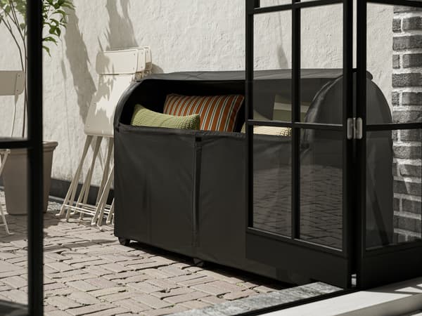 Une boîte de rangement d’extérieur TOSTERÖ noire dans laquelle sont rangés des coussins est posée contre un mur blanc dans un espace extérieur ensoleillé.