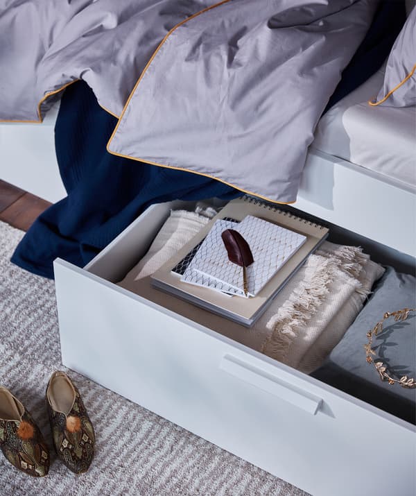 Une boîte de rangement pour lit remplie d’objets dont vous pourriez avoir besoin placée sous le cadre de lit BRIMNES avec rangement et tête de lit.
