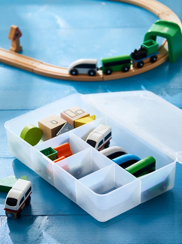 Une boîte GLIS remplie d’accessoires bien rangés, près d’un train miniature et d’une voie ferrée en bois.