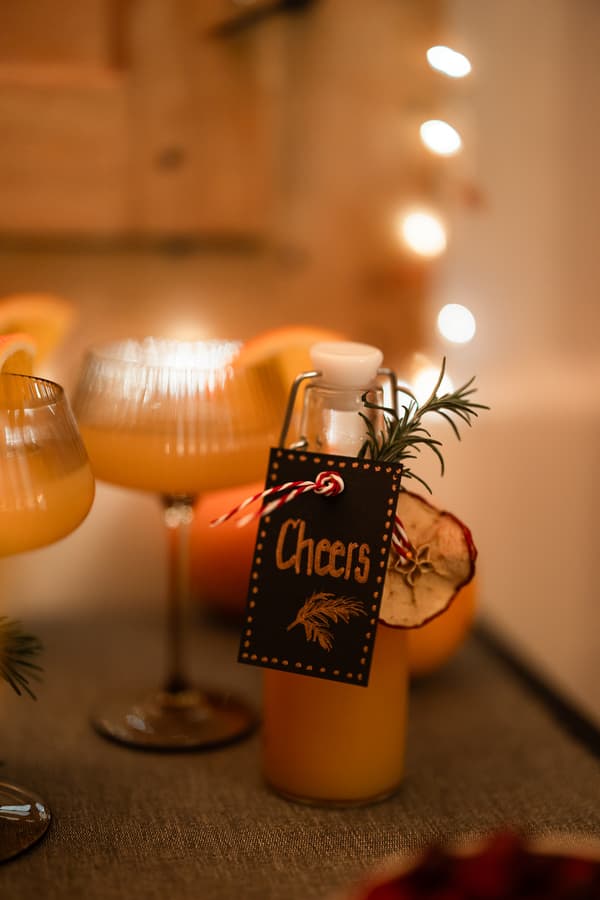Une bouteille en verre avec une étiquette « Cheers » repose parmi des verres à cocktail, garnis de fruits secs et de romarin, sur fond de lumières festives.
