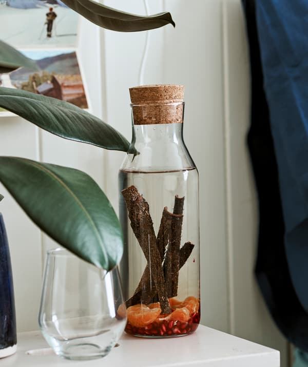Une carafe en verre avec un bouchon en liège remplie d’eau qui infuse avec des bâtons de cannelle et des fruits, à côté d’un verre.