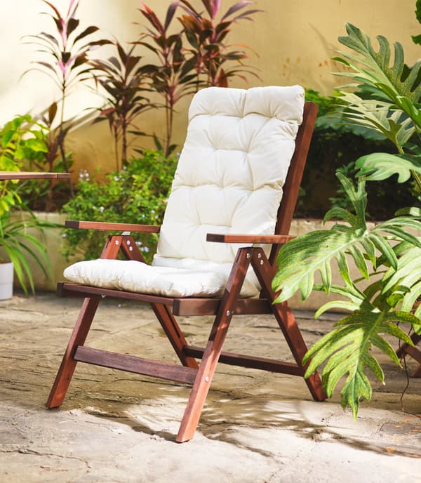 Une chaise à dossier réglable en bois foncé ornée de coussins blancs, placée à l’extérieur et entourée de différentes plantes.